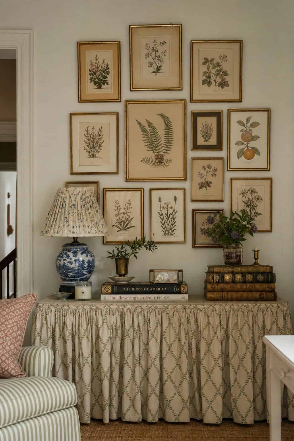 Grandmillennial wall decor idea showing a collected botanical print cluster in antique gold frames above a skirted console, styled with porcelain, old books, and soft daylight. Perfect for Pinterest searches like botanical gallery wall, grandmillennial decor ideas, vintage wall art, and new traditional home decor, this layered look feels classic, cozy, and easy to save for a charming living room or hallway wall.