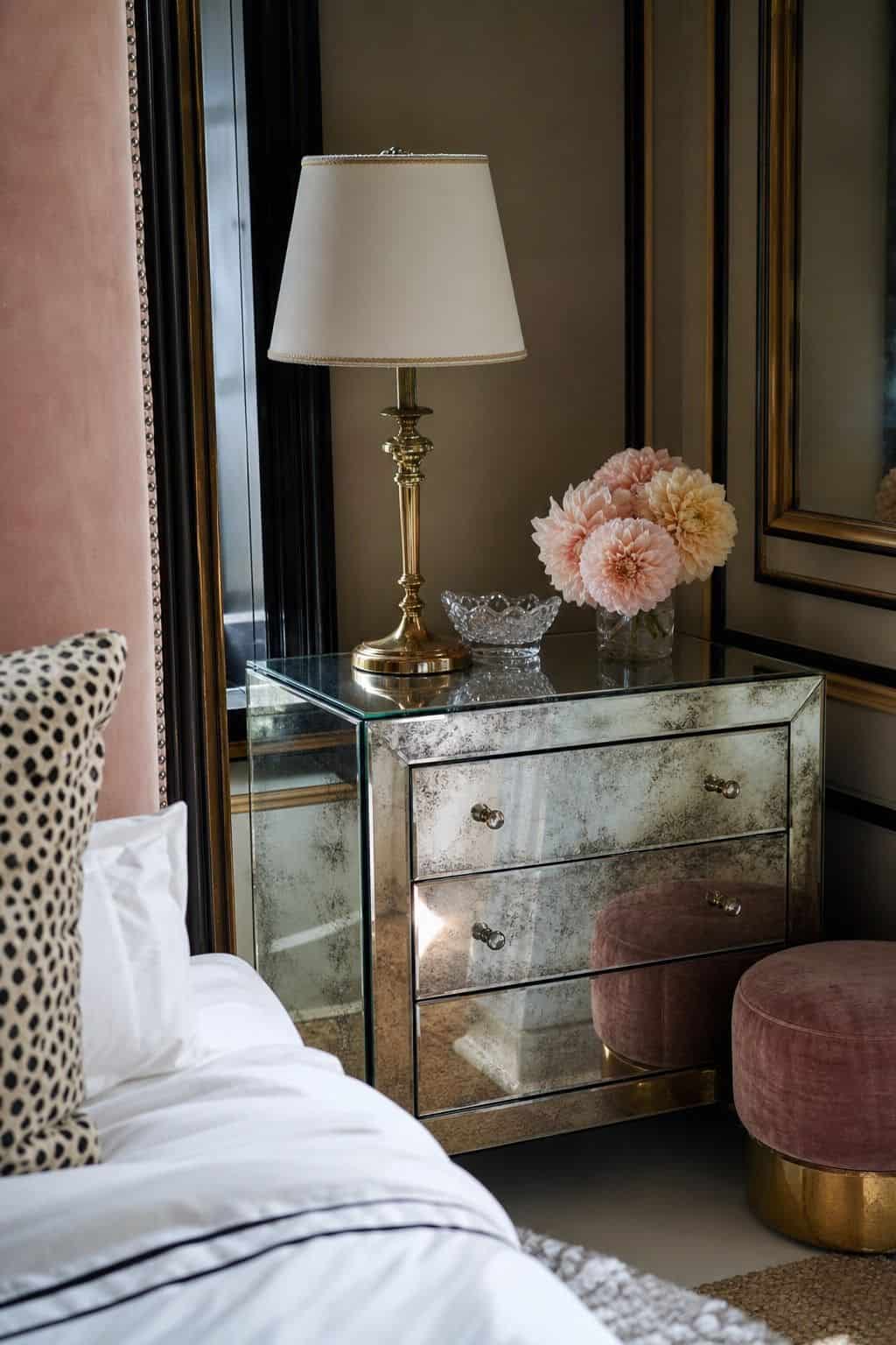 A glamorous Hollywood Regency bedroom corner with a mirrored chest reflecting soft daylight, a gold lamp with an ivory shade, blush flowers, a crystal dish, velvet seating, and polished brass details. The mirrored furniture brings reflection down to furniture level, making the space feel brighter and more luxurious without losing the realistic texture of wood floors, layered textiles, and collected styling.