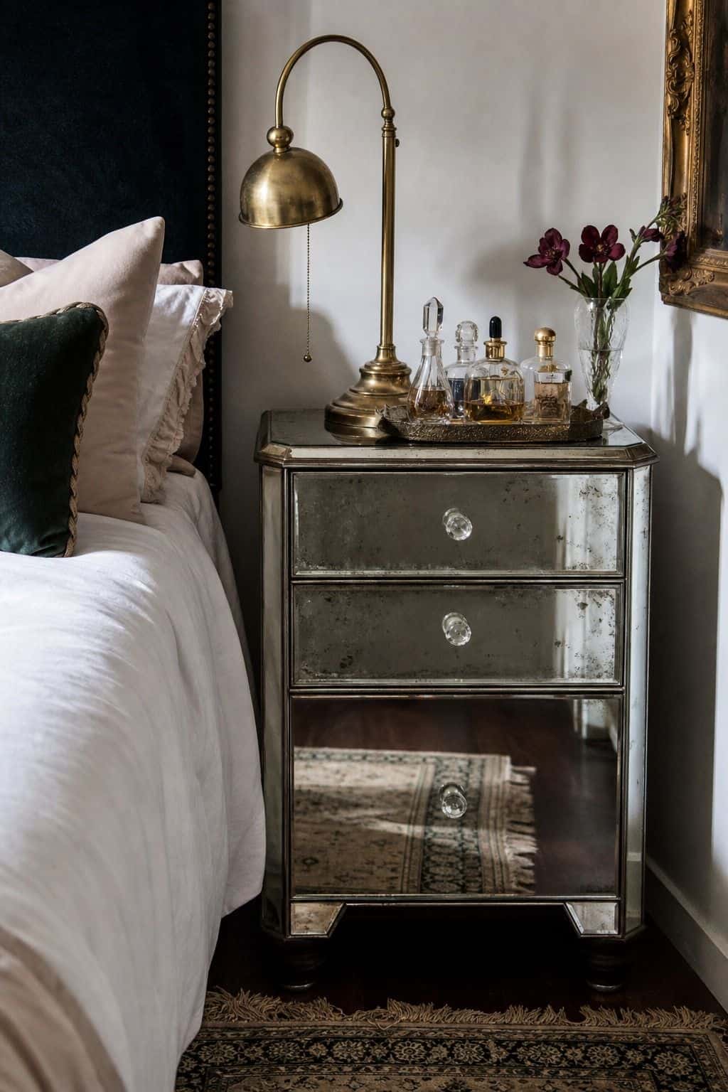 Compact bedroom corner with a secondhand mirrored nightstand, brass lamp, small tray, and vintage perfume bottles without readable labels. Realistic reflections, tiny age marks, and soft daylight make the piece feel found rather than showroom perfect, showing how mirrored furniture can create Hollywood Regency glamour on a secondhand budget. The styling focuses on sparkle, symmetry, and reflection while keeping the room modest enough for a renter or budget decorator to copy.