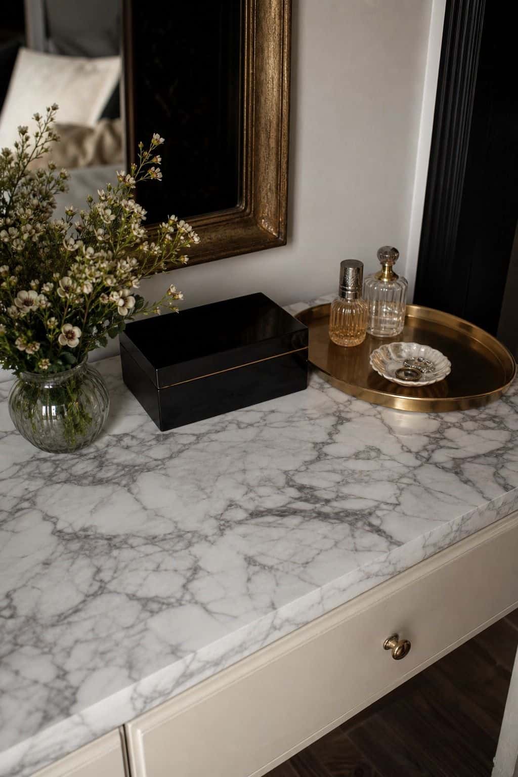 Close view of a budget vanity surface covered in realistic marble contact paper, styled with a gold-rim tray, black lacquer box, and small flowers. Slight seams and believable faux-stone texture keep the project practical, showing how contact paper can create a polished Hollywood Regency surface without the cost or weight of real marble. The scene keeps the project polished but honest, with budget materials elevated through careful styling, contrast, and small metallic details.