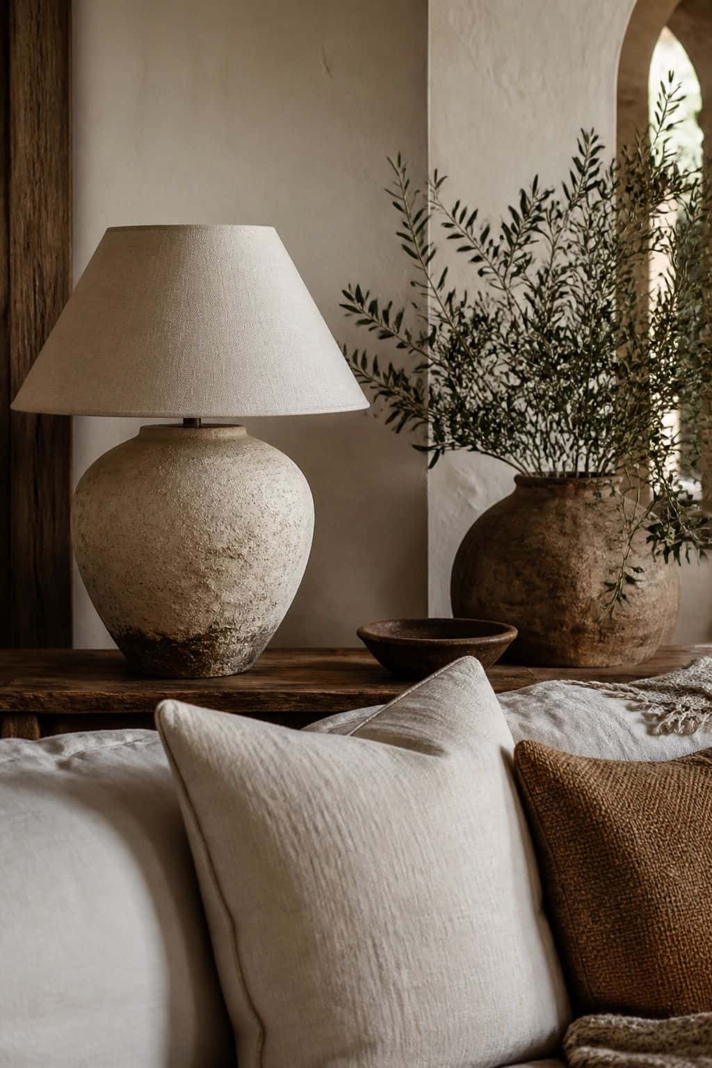 A Ceramic Table Lamp shown in a handmade ceramic table lamp with a linen shade on an aged wood console beside olives, pillows, plaster walls, and relaxed sun-faded Mediterranean textiles. This realistic editorial interior photo is meant for readers saving Mediterranean lighting ideas and warm lighting ideas for a home that feels golden, relaxed, and sun washed. It naturally fits Mediterranean decor ideas, Mediterranean home decor, and warm cozy lighting inspiration without looking staged or.