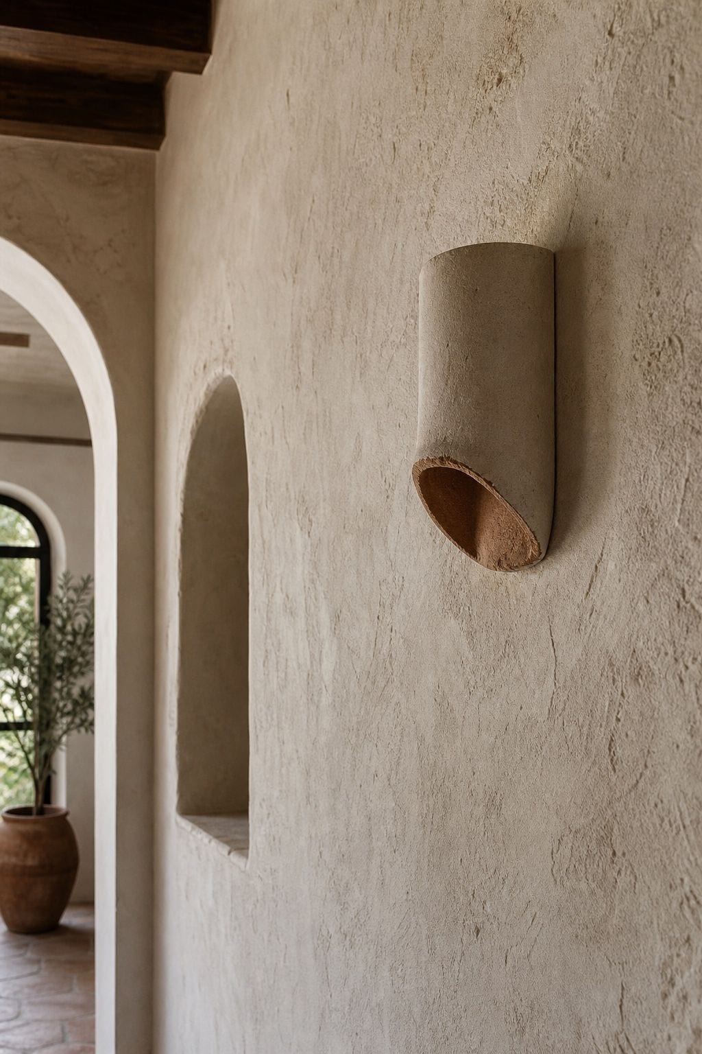 A Plaster or Terracotta Sconce shown in a close plaster hallway wall with a sculptural terracotta sconce, rounded niche, soft archway, and handmade limewash texture in warm earthy tones. This realistic editorial interior photo is meant for readers saving Mediterranean lighting ideas and warm lighting ideas for a home that feels golden, relaxed, and sun washed. It naturally fits Mediterranean decor ideas, Mediterranean home decor, and warm cozy lighting inspiration without looking staged or.