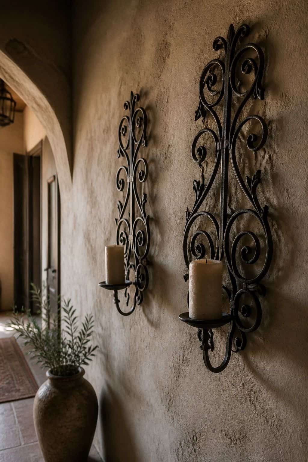 A moody editorial Mediterranean interior photo showing dark wrought-iron candle sconces and wall pieces casting shadows across a warm plaster corridor, styled as realistic inspiration for Wrought-Iron Wall Pieces. The scene uses soft natural daylight, aged plaster, handmade materials, and imperfect lived-in details, making it useful for saving wrought iron wall decor and Mediterranean home decor ideas inspiration. It also fits textured wall decor, warm neutral home decor searches for a warm